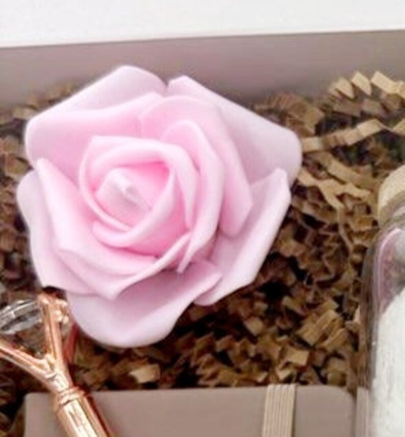May include: A pink rose, a gold-colored pen with a crystal-like top, and a glass jar filled with bath salts. The items are arranged in a white gift box with brown shredded paper.