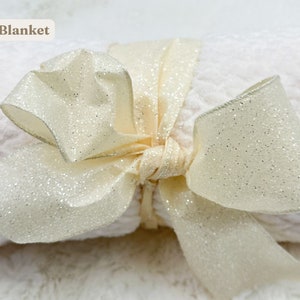 May include: A white blanket with a sparkly ivory ribbon tied in a bow. The blanket is rolled up and the ribbon is tied around it. The text "Blanket" is written in brown text above the blanket.