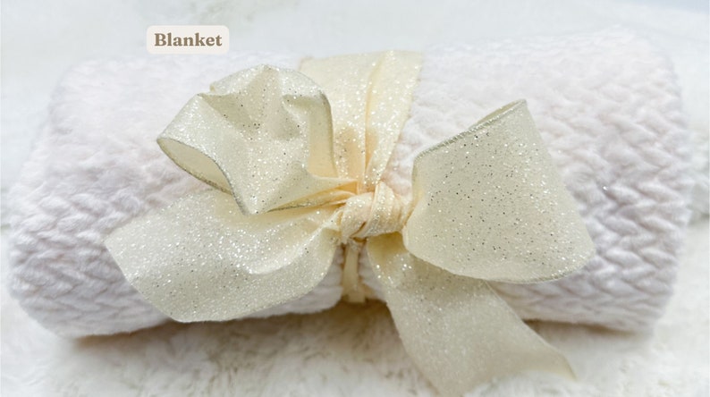 May include: A white blanket with a sparkly ivory ribbon tied in a bow. The blanket is rolled up and the ribbon is tied around it. The text "Blanket" is written in brown text above the blanket.