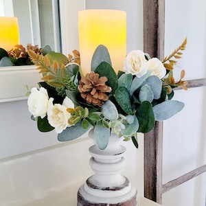 May include: A white pillar candle holder with a brown base, decorated with a faux floral arrangement of white roses, green eucalyptus leaves, and brown pine cones.