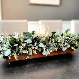 May include: A wooden trough filled with a leafy green floral arrangement. The arrangement includes eucalyptus and small white flowers. A glass candle holder with a lit candle sits in the center of the arrangement.