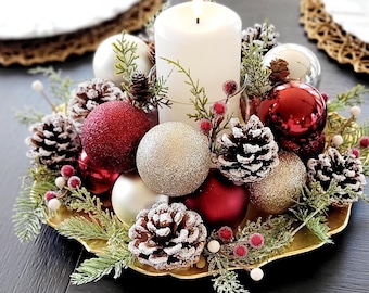 May include: A centerpiece featuring a white pillar candle surrounded by red and gold glittered ornaments, frosted pinecones, green cedar sprigs, and small red and white berries, all arranged on a scalloped gold tray on a dark surface.