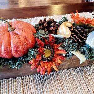 May include: A wooden box centerpiece with a variety of fall decor, including a large orange pumpkin, small white and gold gourds, pine cones, a sunflower, and white flowers.