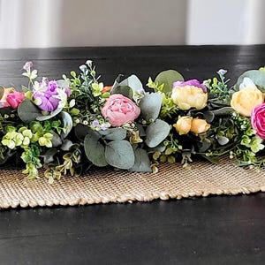 Puede incluir: Un camino de mesa floral decorativo con una variedad de flores artificiales coloridas, incluyendo flores naranjas, amarillas, rosas y moradas, intercaladas con follaje verde. El camino se coloca sobre un mantel individual de yute tejido.