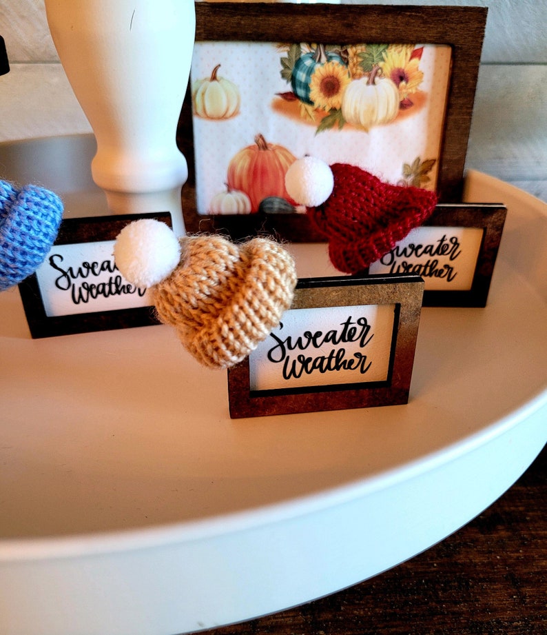 Puede incluir: Tres peque&ntilde;as se&ntilde;ales de madera con el texto "sweater weather" escrito en cursiva negra. Cada se&ntilde;al tiene un peque&ntilde;o gorro de punto en la parte superior, uno azul, uno marr&oacute;n y uno rojo.
