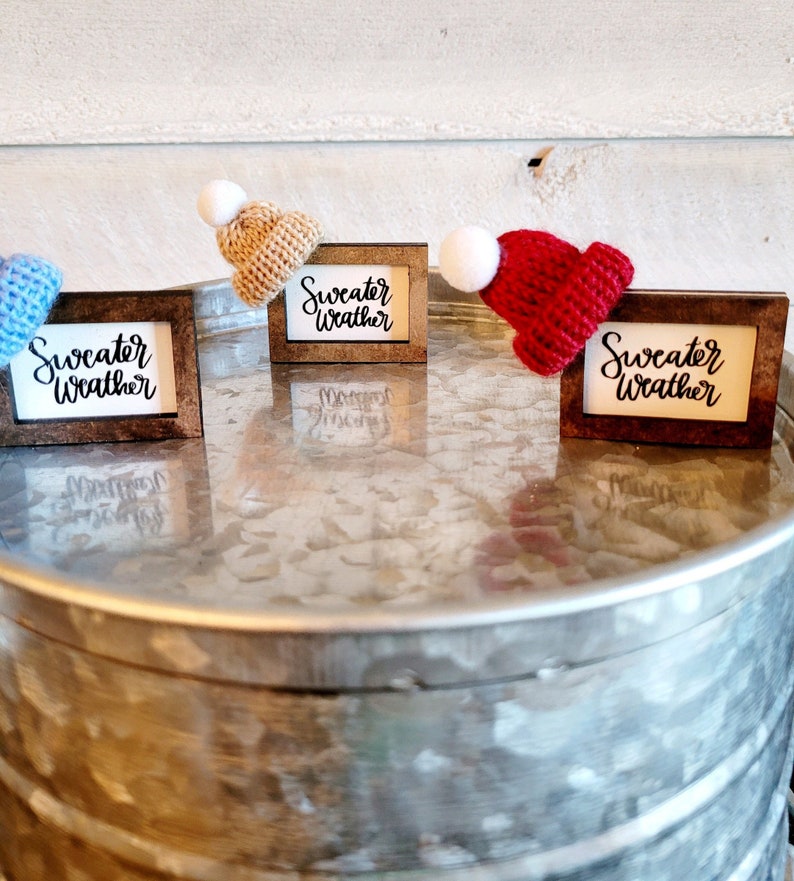 Puede incluir: Tres peque&ntilde;as se&ntilde;ales de madera con el texto "Sweater Weather" en ellas. Cada se&ntilde;al tiene un gorro de punto de un color diferente en la parte superior: azul, tostado y rojo.