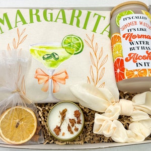May include: A gift box filled with items for a margarita themed party. The box includes a white tote bag with the word "Margarita" in green letters, a glass jar with a label that says "It's called Summer Water. It's like Normal Water but has Alcohol in it", a small white candle, a dried orange slice, and a white scrunchie.