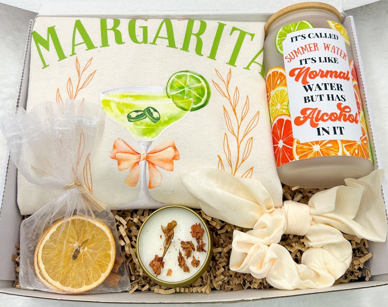 May include: A gift box filled with items for a margarita themed party. The box includes a white tote bag with the word "Margarita" in green letters, a glass jar with a label that says "It's called Summer Water. It's like Normal Water but has Alcohol in it", a small white candle, a dried orange slice, and a white scrunchie.