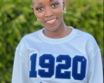 Op de afbeelding: Een vrouw draagt een wit sweatshirt met een blauwe geborduurde patch waarop "1920" en "Zeta Phi Beta Sorority, Incorporated" staat.