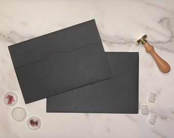 May include: Two black envelopes with a flap on the top, a wooden handle with a gold wax seal, and three white wax seals with dried flowers on a white marble surface.