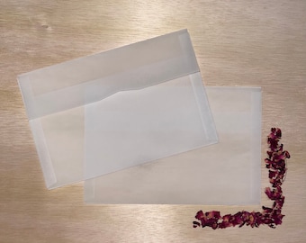 May include: Two translucent white envelopes are arranged on a light wood surface, with one partially overlapping the other. A small arrangement of dried, dark red rose petals is in the lower right corner. The envelopes have a simple, clean design.