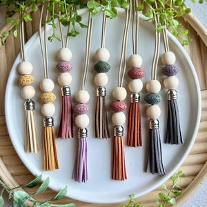 May include: A collection of aromatherapy car diffusers with wooden beads, lava stones, and colorful leather tassels. The diffusers are displayed on a white plate, with a neutral background and greenery.