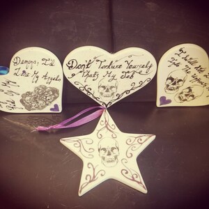May include: Four white ceramic ornaments with dark text and skull designs. Three are heart-shaped, and one is star-shaped. The hearts have phrases like "Demons, I'd lose my angels" and "Don't torture yourself, that's my job." Each has a ribbon for hanging.