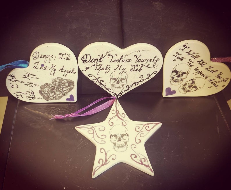 May include: Four white ceramic ornaments with dark text and skull designs. Three are heart-shaped, and one is star-shaped. The hearts have phrases like "Demons, I'd lose my angels" and "Don't torture yourself, that's my job." Each has a ribbon for hanging.