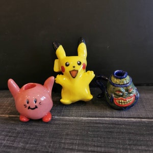May include: Three ceramic figurines: a pink Kirby-like character, a yellow Pikachu, and a green and blue monster-faced teapot. The figurines are on a dark wood-grain surface.