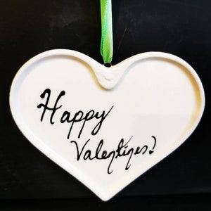 May include: White heart-shaped ornament with the words "Happy Valentine's!" written in black script. A green ribbon is attached for hanging. The ornament is likely ceramic or porcelain.