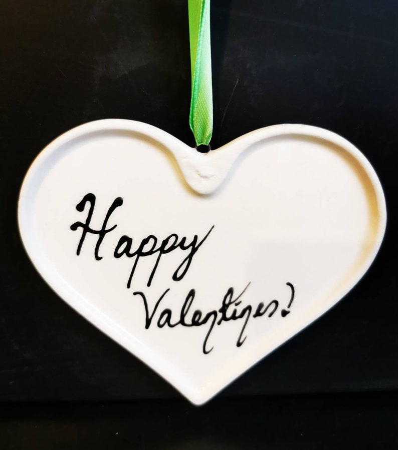 May include: White heart-shaped ornament with the words "Happy Valentine's!" written in black script. A green ribbon is attached for hanging. The ornament is likely ceramic or porcelain.