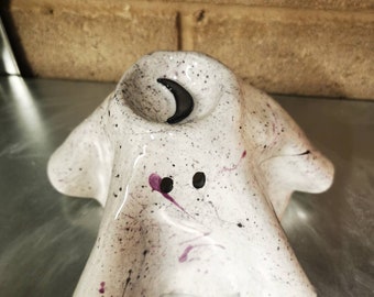 May include: A white ceramic ghost-shaped candle holder with black and purple splatter paint. The ghost has two black eyes and a black crescent moon inside the candle holder. The candle holder is on a metallic surface.