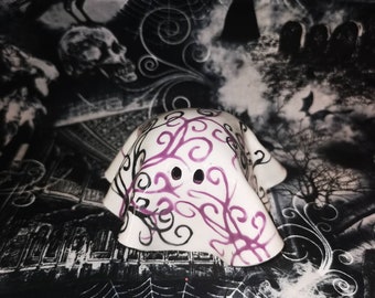 May include: A white ceramic ghost figurine with purple and black swirl designs and two black eye holes. The ghost is set against a backdrop of a Halloween-themed fabric with spiders, webs, and a crow.