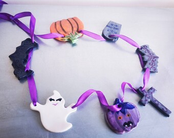 Puede incluir: Una guirnalda de Halloween con amuletos de cerámica en forma de calabaza, fantasma, murciélago y cruz, ensartados en una cinta morada. La calabaza es naranja con detalles verdes, y el fantasma es blanco con pestañas negras. Ideal para la decoración festiva.