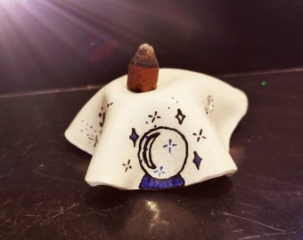 May include: A white ceramic incense holder with a hand-painted design featuring a crystal ball, crescent moon, and stars. A brown incense cone sits atop the holder. The holder is on a dark surface.