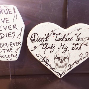 May include: Two white, hand-painted decorative pieces. One is coffin-shaped with the text "TRUE LOVE NEVER DIES" and "2019-EVER THE HOLDENS". The other is heart-shaped with a skull and the text "Don't Torture Yourself That's My Job".
