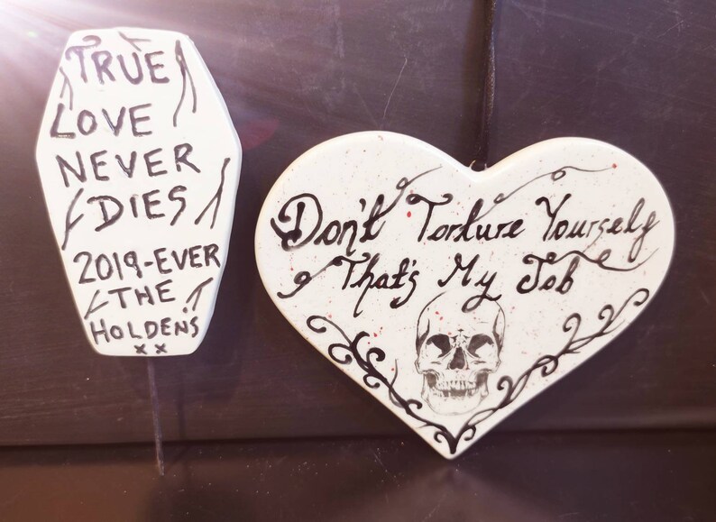 May include: Two white, hand-painted decorative pieces. One is coffin-shaped with the text "TRUE LOVE NEVER DIES" and "2019-EVER THE HOLDENS". The other is heart-shaped with a skull and the text "Don't Torture Yourself That's My Job".