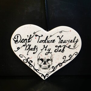 May include: White heart-shaped decorative sign with the text "Don't Torture Yourself That's My Job" in black script. A skull graphic is centered below the text, with black decorative flourishes. Red speckles add detail.