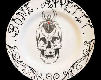 Puede incluir: Plato de cerámica blanca con un diseño de calavera y araña negras. Las palabras "BONE APPETIT" están escritas en una fuente decorativa alrededor del borde. También hay adornos decorativos negros.
