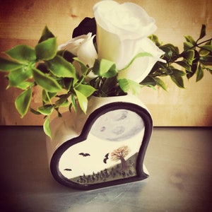 Può includere: Un vaso in ceramica a forma di cuore con bordo nero, raffigurante una scena spettrale con luna piena, pipistrelli e un cimitero. Il vaso contiene rose bianche e fogliame verde.