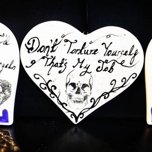 May include: Three heart-shaped ornaments with handwritten gothic text and skull illustrations. The ornaments are white, with blue and purple accents. The text includes phrases like "Demons, I'd lose my angels" and "Don't torture yourself & that's my job."
