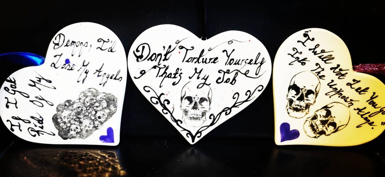 May include: Three heart-shaped ornaments with handwritten gothic text and skull illustrations. The ornaments are white, with blue and purple accents. The text includes phrases like "Demons, I'd lose my angels" and "Don't torture yourself & that's my job."