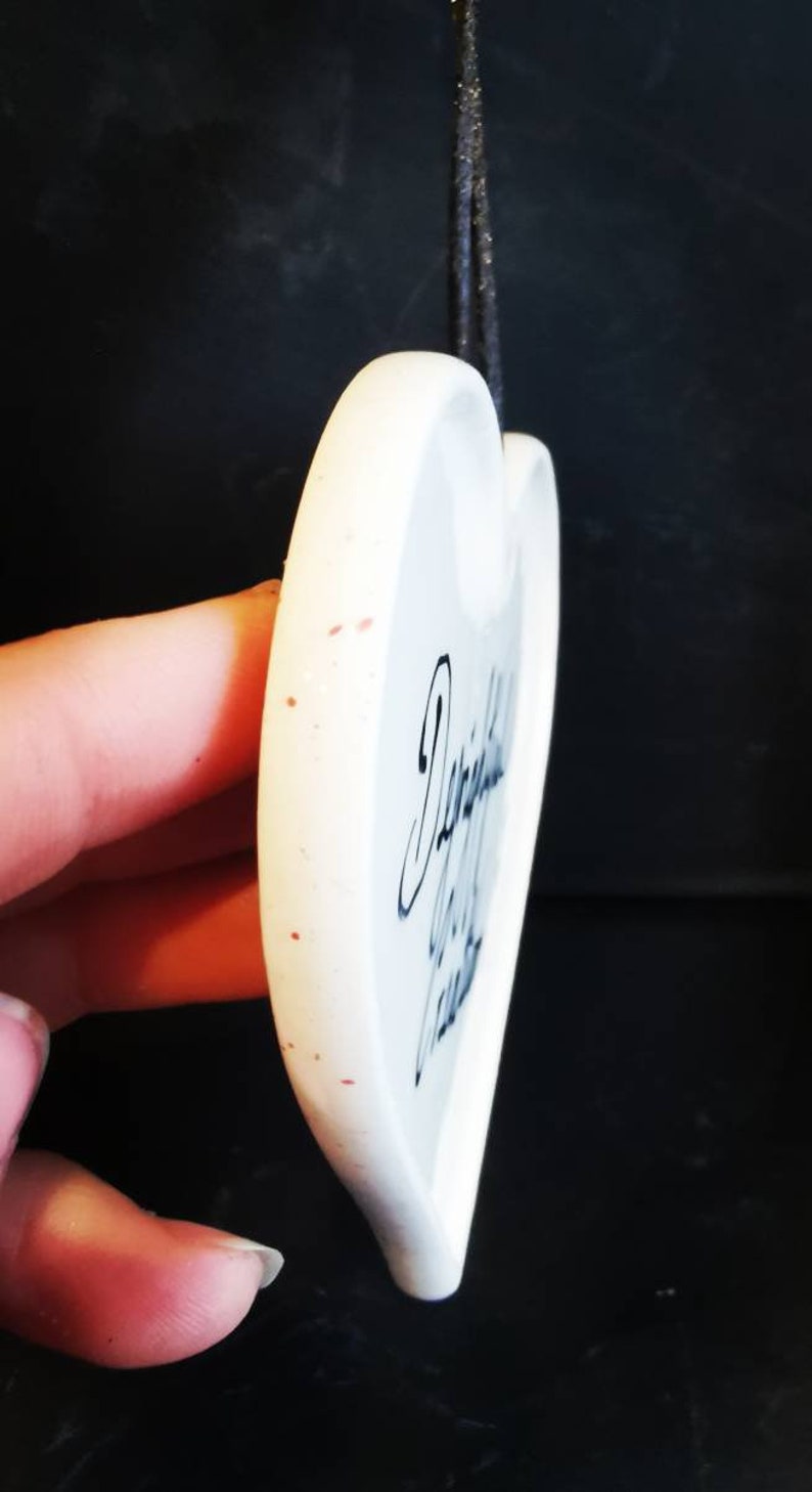 May include: A white heart-shaped ornament with black script. The ornament has a glossy finish and a small hole at the top for hanging. Red speckles dot the surface. A black string is attached for hanging.