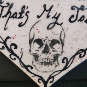May include: A white, diamond-shaped decorative item with the phrase "That's My Job" in black script. A detailed skull illustration is centered below the text. The design includes black flourishes and red speckles.