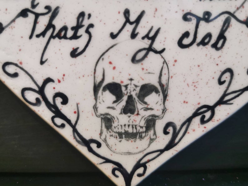 May include: A white, diamond-shaped decorative item with the phrase "That's My Job" in black script. A detailed skull illustration is centered below the text. The design includes black flourishes and red speckles.