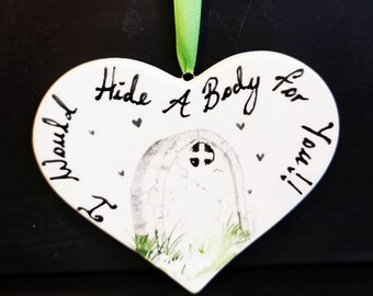 May include: White heart-shaped ornament with the text "I Would Hide A Body For You!" in black script. A tombstone illustration is in the center, with a green ribbon for hanging. Small black hearts are scattered around the text.