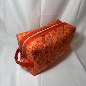 May include: An orange zippered pouch with a floral pattern in shades of orange and red. The pouch has a rectangular shape with a handle on one side and a silver zipper. The background is a white surface.