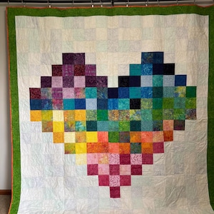Puede incluir: Un edredón hecho a mano con un diseño de corazón hecho de parches cuadrados coloridos. La forma del corazón está compuesta por varios tonos de morado, azul, verde, amarillo, naranja y rosa. El edredón tiene un fondo blanco con un borde verde.