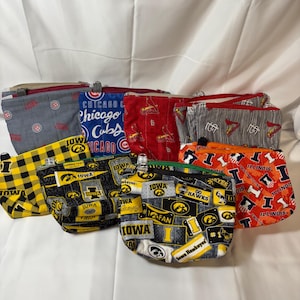 May include: A collection of fabric zipper pouches in various sizes and patterns. Designs include Chicago Cubs logos, Iowa Hawkeyes, and Illinois Fighting Illini. The pouches are in shades of red, blue, black, yellow, and orange.