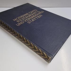 May include: A dark blue hardback book with gold lettering on the spine that reads "Moderne Cafes Restaurants und Vergnugungsstatten".
