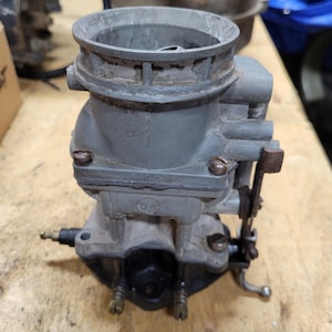 May include: A gray metal automotive carburetor featuring a cylindrical air intake at the top, a central body with visible screws, and a black base section with small metal nozzles and a mechanical linkage arm attached to the side of the unit.
