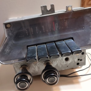 May include: Vintage car radio with a black face and silver trim. The radio has a dial with numbers 5 to 16 and a red needle. There are five black buttons below the dial and two black knobs below the buttons.