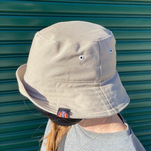 May include: A beige bucket hat with a black Dickies logo on the side. The hat has two white eyelets on the top.