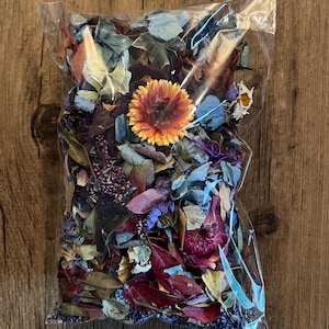 Peut inclure: Un sac en plastique transparent rempli de pétales de fleurs séchées et de feuilles dans des tons de bleu, violet, rouge, orange et jaune. Le sac est sur une surface en bois.