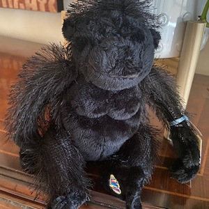 May include: Black plush gorilla toy with a colorful tag attached to its wrist. The gorilla has a shaggy black fur and a friendly expression.
