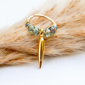 May include: A gold hoop earring featuring a central gold spike charm and four small, translucent, iridescent green beads arranged on either side of the charm. The hoop is constructed from thin gold wire, creating a delicate and circular structure.