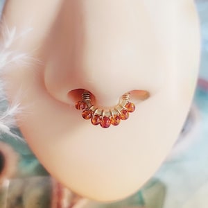 Puede incluir: Primer plano de una nariz con un anillo de tabique. El anillo es de color dorado y presenta una serie de pequeñas cuentas de color rojo anaranjado. La nariz se encuentra sobre un fondo borroso con iluminación suave.