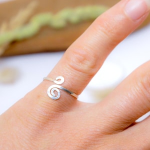 Puede incluir: Un anillo plateado con un diseño en espiral se lleva en un dedo. El anillo tiene un estilo sencillo y elegante. El anillo es una joya. La marca "Melle Saraëlle" está en la esquina superior izquierda.