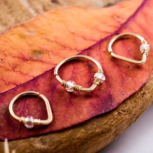 May include: Three gold-colored hoop earrings with small, clear beads are displayed on a vibrant red and orange leaf. The earrings are arranged on a piece of wood, with a dried plant in the background.