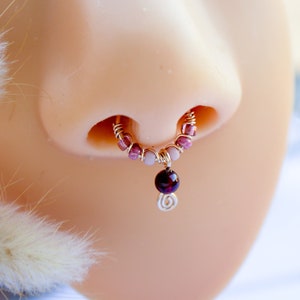 Puede incluir: Un aro de nariz dorado con una serie de pequeñas cuentas rosas y una cuenta roja oscura colgante. El aro de nariz está diseñado para ser usado en el tabique nasal. La joya se muestra en una cabeza de maniquí.